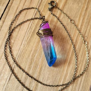 Puede incluir: Un collar con colgante de cristal colorido. El cristal, que va del rosa al azul, está envuelto en alambre dorado. La cadena dorada descansa sobre una superficie de madera.