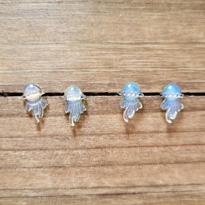 May include: Four iridescent blue and white jellyfish earrings on a wooden surface. The earrings are small and delicate, with a clear resin finish.