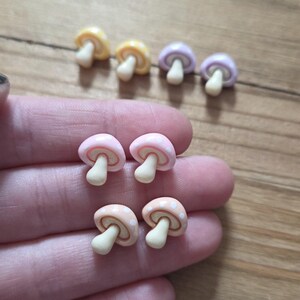 May include: A collection of miniature mushroom-shaped charms in various pastel colors: yellow, lavender, pink, and peach. Each mushroom has a white stem and a dotted cap. These small charms are likely for crafting or jewelry making.