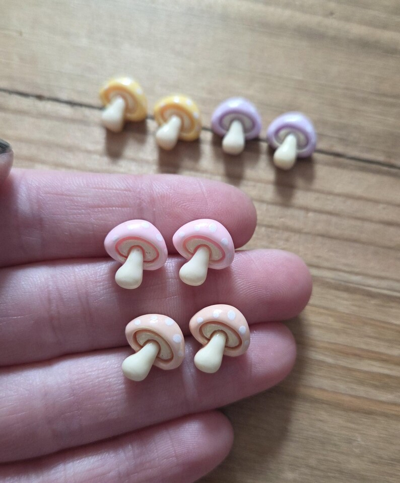 May include: A collection of miniature mushroom-shaped charms in various pastel colors: yellow, lavender, pink, and peach. Each mushroom has a white stem and a dotted cap. These small charms are likely for crafting or jewelry making.