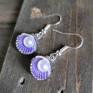 May include: A pair of seashell earrings. Each earring features a lavender-colored shell with a silver-colored trim and a white pearl in the center. The earrings have silver-colored fish hook ear wires.