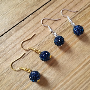 May include: Four pairs of dangle earrings with dark blue, sparkly, round beads. Two pairs have gold-colored hooks, and two pairs have silver-colored hooks. The earrings are displayed on a wooden surface.