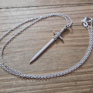 Puede incluir: Un collar con colgante plateado con una espada en miniatura. La espada tiene detalles intrincados en la empuñadura y una hoja puntiaguda. La cadena es de diseño delicado y enlazado. La joya se exhibe sobre una superficie de madera.