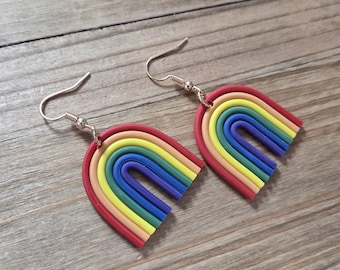 Puede incluir: Un par de pendientes en forma de arcoíris. Cada pendiente presenta un arco rojo con capas de amarillo, naranja, verde, azul y morado. Los pendientes tienen ganchos plateados.