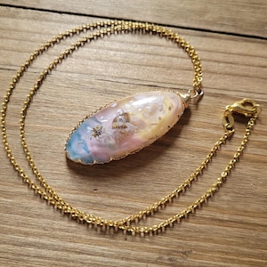 May include: A gold chain necklace with a large oval pendant. The pendant is made of a translucent material with a pink, blue, and white swirl pattern. The pendant is surrounded by a gold wire frame.