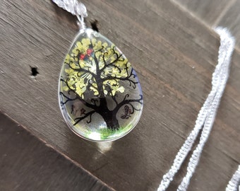 Puede incluir: Un collar de cadena plateada con un colgante en forma de lágrima que presenta un árbol negro con flores amarillas y rojas dentro de una resina transparente.