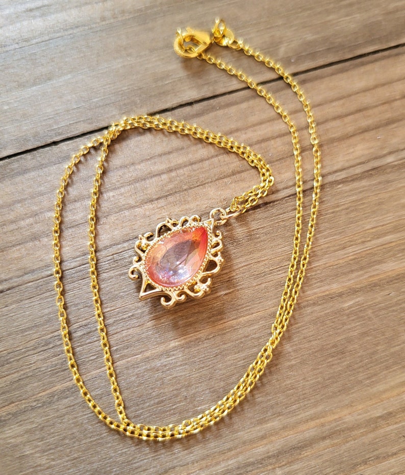 May include: A gold chain necklace with a teardrop-shaped pendant featuring a pink and orange gem set in a gold filigree frame.