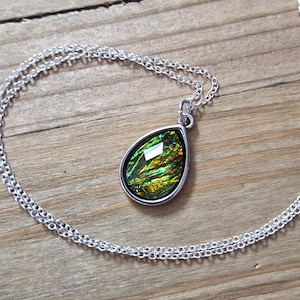 May include: Silver-toned necklace with a teardrop pendant. The pendant features a vibrant, iridescent gemstone with shades of green, yellow, and gold. The chain is a delicate double strand, resting on a wooden surface.