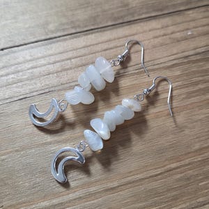 Puede incluir: Un par de pendientes colgantes con un dije de media luna plateada y una hebra de cuentas claras de forma irregular. Los pendientes tienen ganchos para la oreja plateados. Los pendientes se exhiben sobre una superficie de madera.