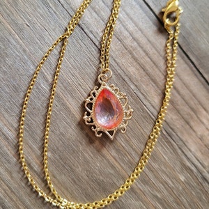 May include: A gold chain necklace with a teardrop-shaped pendant featuring a pink and orange gem. The pendant has a delicate, ornate design.