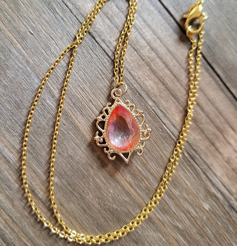 May include: A gold chain necklace with a teardrop-shaped pendant featuring a pink and orange gem. The pendant has a delicate, ornate design.