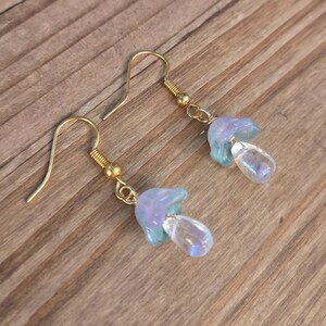 May include: A pair of gold-coloured hook earrings. Each earring features a light blue and purple flower-shaped charm with a clear, teardrop-shaped bead. The earrings are displayed on a wooden surface.
