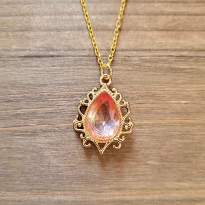May include: A gold chain necklace with a pink teardrop-shaped pendant. The pendant is set in a gold filigree design.