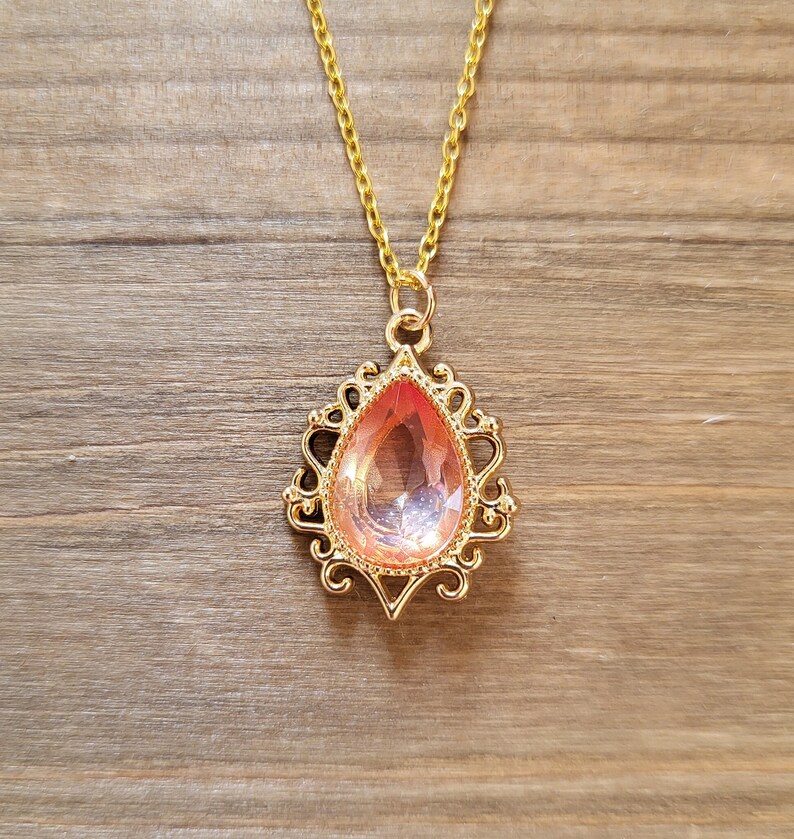 May include: A gold chain necklace with a pink teardrop-shaped pendant. The pendant is set in a gold filigree design.
