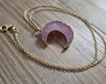 May include: A gold-colored chain necklace with a crescent moon pendant. The pendant has a light purple, crystal-like texture with gold-colored edges. The necklace is displayed on a wooden surface.