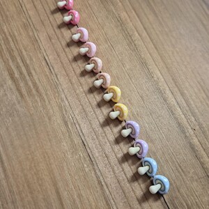 May include: A diagonal row of colorful mushroom-shaped beads. The beads are in shades of pink, yellow, purple, and blue, with white stems. Some have polka dot patterns. The beads are arranged on a wooden surface.