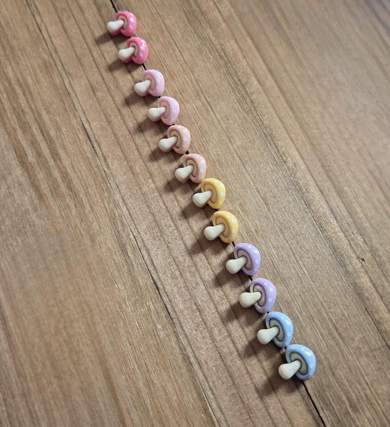 May include: A diagonal row of colorful mushroom-shaped beads. The beads are in shades of pink, yellow, purple, and blue, with white stems. Some have polka dot patterns. The beads are arranged on a wooden surface.