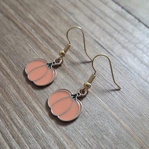May include: A pair of pumpkin-shaped earrings. Each earring features a light orange pumpkin charm with a green stem and gold-colored ear wires. The earrings are displayed on a wooden surface.