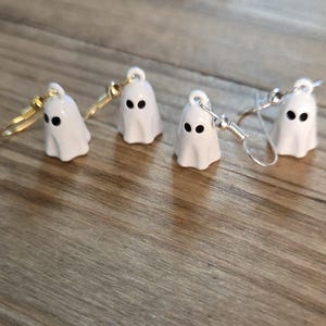 Pode incluir: Quatro brincos de fantasma brancos com olhos pretos e ganchos de brinco dourados e prateados. Os brincos são pequenos, com um design simples, e estão colocados sobre um fundo de madeira. Um acessório divertido para o Halloween.