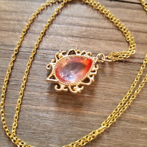 May include: A gold chain necklace with a teardrop-shaped pendant featuring a pink and orange iridescent glass stone set in a gold filigree frame.