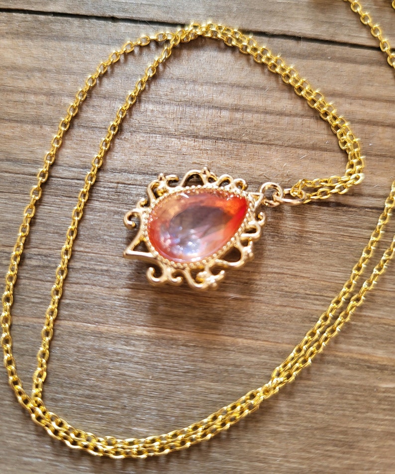May include: A gold chain necklace with a teardrop-shaped pendant featuring a pink and orange iridescent glass stone set in a gold filigree frame.