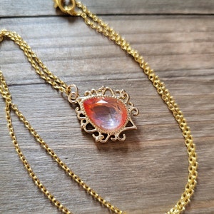 May include: A gold chain necklace with a teardrop-shaped pendant featuring a large, pink, iridescent gemstone set in a gold filigree frame.