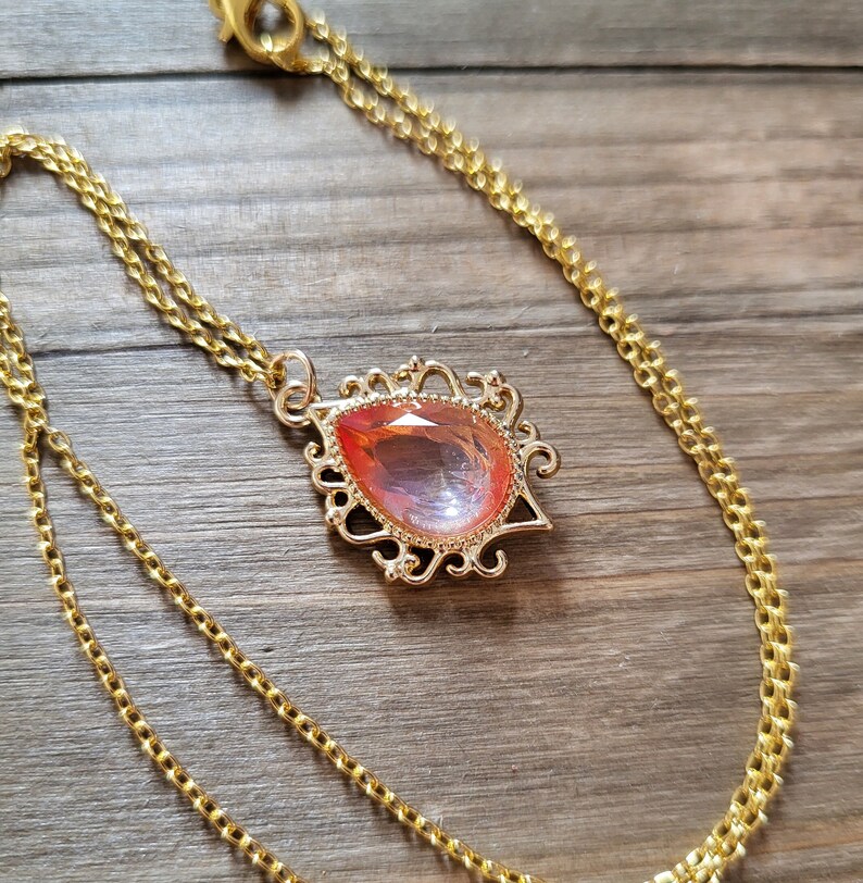 May include: A gold chain necklace with a teardrop-shaped pendant featuring a large, pink, iridescent gemstone set in a gold filigree frame.