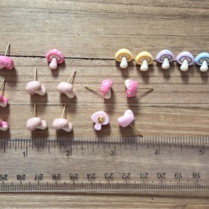 May include: A collection of mushroom-shaped stud earrings in various colors, including pink, yellow, purple, and blue. The earrings are arranged on a wooden surface next to a ruler marked in inches. The earrings have gold-colored posts.
