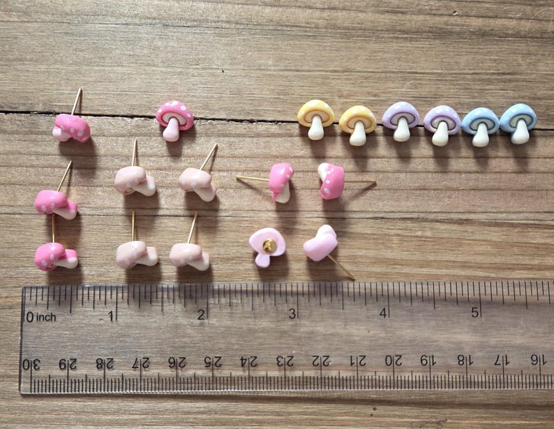 May include: A collection of mushroom-shaped stud earrings in various colors, including pink, yellow, purple, and blue. The earrings are arranged on a wooden surface next to a ruler marked in inches. The earrings have gold-colored posts.