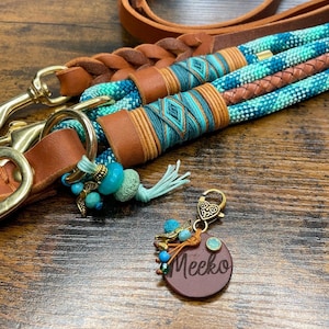 Könnte beinhalten: Eine braune Lederhundeleine mit geflochtenem Design und türkisfarbenen und blauen Akzenten aus geflochtenem Seil. Ein passendes Leder-Hundehalsband mit dem Namen "Meeko" ist mit einem goldenen Anhänger an der Leine befestigt.