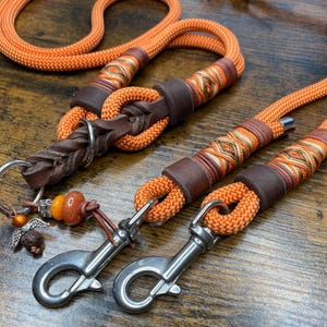 Könnte beinhalten: Orangefarbene Hundeleine aus Seil mit braunen Lederakzenten und silberfarbenen Beschlägen. Die Leine hat dekorative Nähte und eine geflochtene Lederschlaufe. Ein kleiner Anhänger mit einer geflügelten Figur und Perlen ist befestigt.