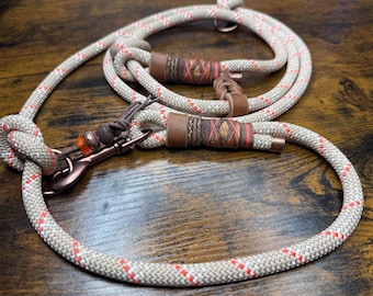 May include: A beige and red rope dog leash with copper-colored hardware. The leash features decorative brown leather and woven accents. The leash is knotted at one end and has a clasp for attaching to a dog collar.