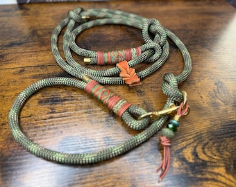 May include: A dog leash made of green and brown camouflage rope. The leash has decorative orange and green wrapping and a gold-colored clasp. A small orange leaf-shaped charm is attached to the leash.