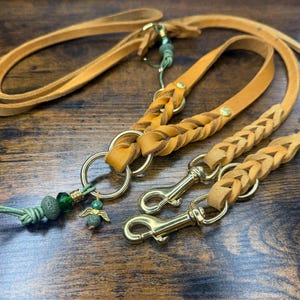 May include: A tan leather dog lead with a braided section and gold-coloured hardware. The lead features a decorative charm with green beads and a small angel pendant. Designed for pet owners.