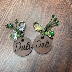May include: Two brown leather keychains with the name "Dali" engraved on them. Each keychain has a silver or gold heart-shaped clasp and a green beaded tassel with a small angel charm.