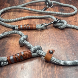 以下が含まれることがあります： 茶色の革のアクセントと銀色の金属製のクリップが付いた灰色のロープ製の犬のリード。リードは結び目があり、ハンドルが付いています。