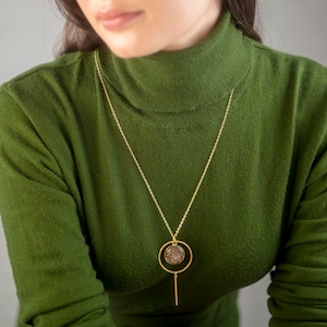 Peut inclure: Collier doré avec un pendentif circulaire orné d'un motif brun texturé. Le pendentif est suspendu à une chaîne délicate, avec une barre verticale en dessous. Le collier est porté sur un col roulé vert.