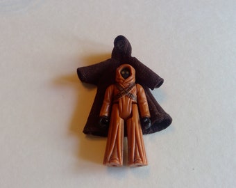 May include: A brown plastic action figure of a Star Wars character wearing a brown hooded robe. The figure has a black face mask and is holding a brown weapon.
