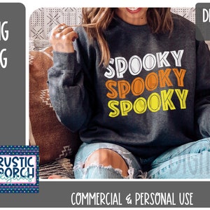 Puede incluir: Una sudadera gris con el texto "Spooky" repetido tres veces en un diseño superpuesto. El texto está en blanco, naranja y amarillo. La sudadera la lleva puesta una persona sentada en un sofá.