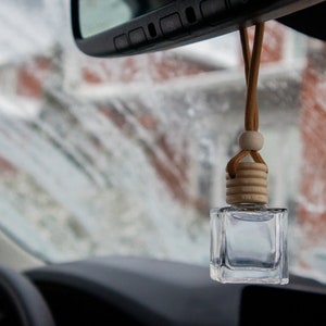Puede incluir: Un frasco de vidrio transparente pequeño y cuadrado que cuelga del espejo retrovisor de un coche mediante un cordón color canela. El frasco tiene un tapón de madera clara con estrías horizontales y una cuenta de madera redonda.