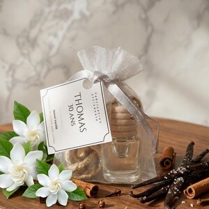 Peut inclure: Un diffuseur de parfum pour voiture en verre transparent avec un bouchon en bois est à l'intérieur d'un sac blanc transparent noué avec un ruban. Une étiquette indique "THOMAS 30 ANS". Gousses de vanille, bâtons de cannelle et fleurs blanches.