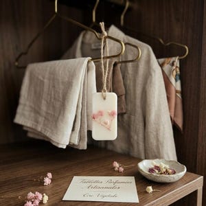 Puede incluir: Una tableta de cera rectangular blanquecina con flores secas rosas cuelga de una cuerda de yute. La tableta está suspendida frente a una camisa y pantalones de lino beige colgados en un armario de madera. Un pequeño cuenco de cerámica y una tarjeta con texto en francés están en el estante.