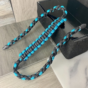 May include: A black and turquoise braided paracord strap with a loop at one end. The strap is made of two different types of paracord, one black and one turquoise, that are braided together.