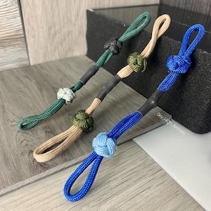 May include: Four different colored rope lanyards with knots at the end. The lanyards are green, blue, tan, and black. The knots are all different colors and patterns. The lanyards are all tied to a black surface with the text "Sky High Archery" printed on it.