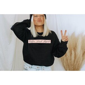 Op de afbeelding: Een zwart sweatshirt met de tekst "Coffee, Teach, Repeat" in roze schrift op een roze banner.