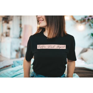 Puede incluir: Una camiseta negra con el texto "Coffee. Teach. Repeat." impreso en rosa sobre una pancarta rosa claro.