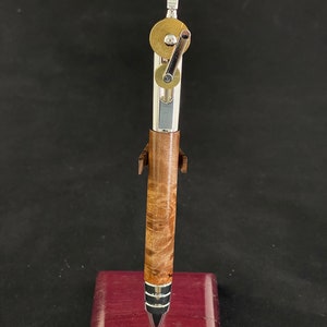 May include: A silver and wood pen with a brass mechanism and a silver tip. The pen is on a burgundy wood stand.