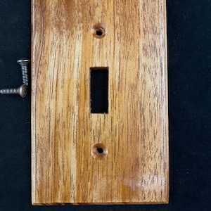 Puede incluir: Una tapa de interruptor de luz de madera con un acabado de veta de madera natural. La tapa tiene una forma rectangular con un recorte para un interruptor de luz.