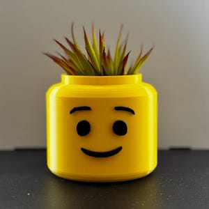 May include: A bright yellow planter shaped like a Lego head with a smiling face. The planter is filled with green and red-tipped faux succulents. The planter is sitting on a black surface.