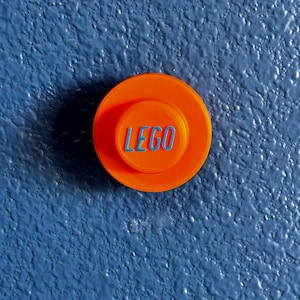 May include: Close-up of an orange LEGO brick with the word "LEGO" in blue lettering. The brick is circular and sits against a textured blue background.